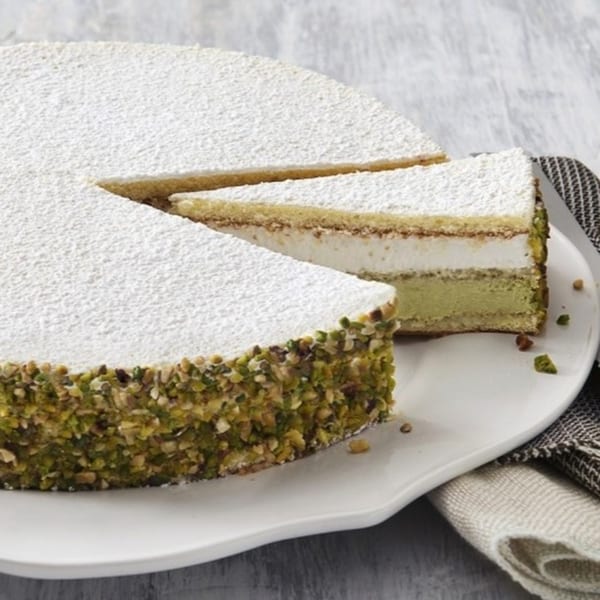 A round pistachio cake with a slice removed, dusted with powdered sugar on a white plate.